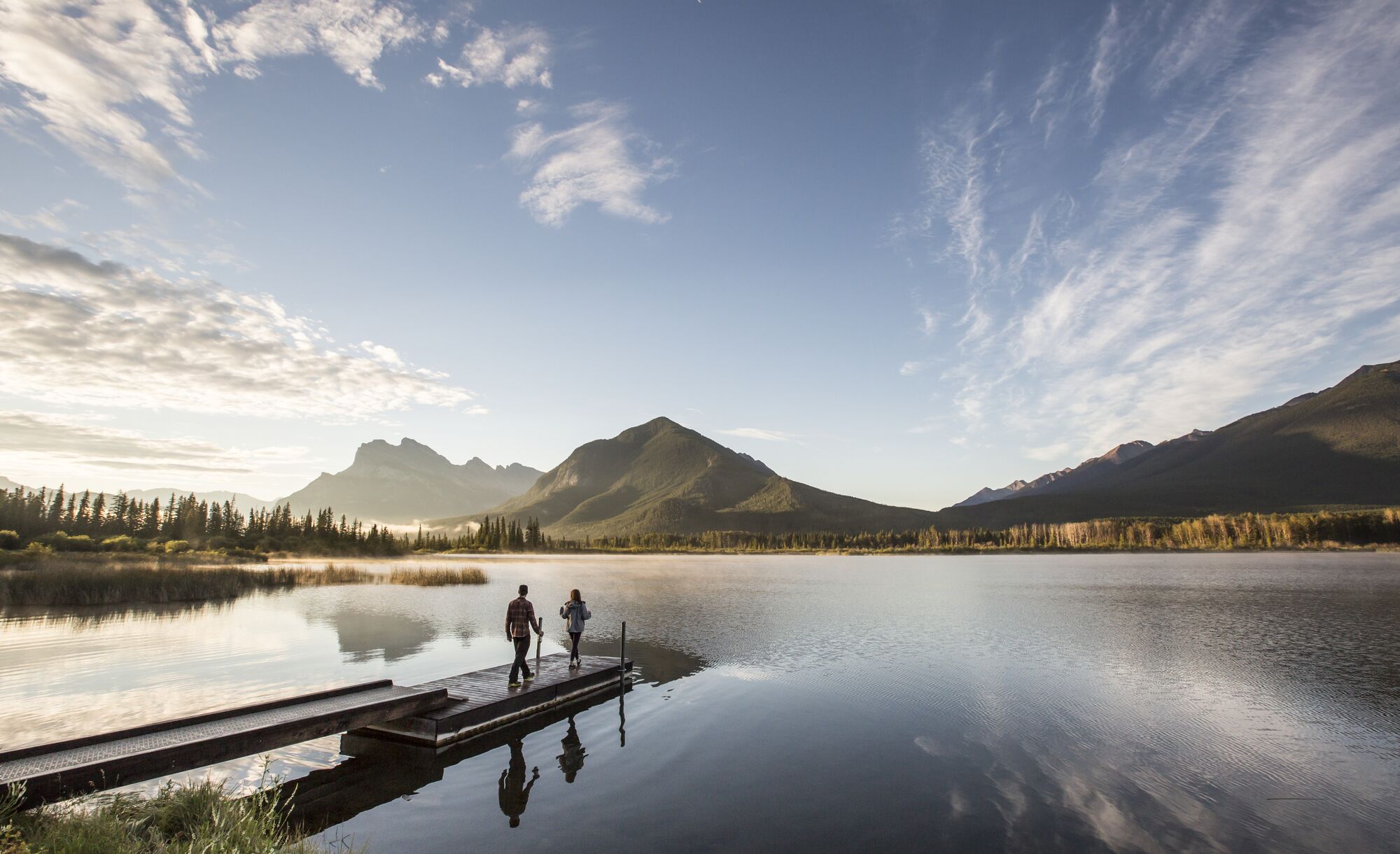 A couple walks along a dock towards a Vermilion Lake framed by Mount Rundle and Sulphur Mountain in Banff National Park.